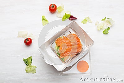 Top view sandwich with soft cheese and red fish lies in the lunch box next to the greens and tomatoes on a white table Stock Photo