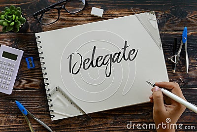 Top view of stationery glasses, calculator,plant and hand holding pen writing Delegate on notebook on wooden background Stock Photo