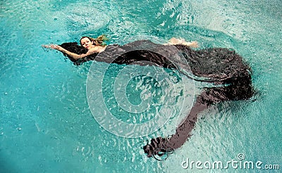 Top view to a beautiful sexy young woman floating like a mermaid in black cloth dress wrapped in green water of the swimming pool Stock Photo