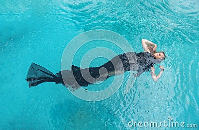 Top view to a beautiful sexy young woman floating like a mermaid fish, wrapped in black cloth dress in turquoise blue water of the Stock Photo