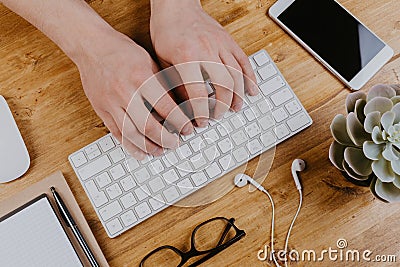 Top View of trendy wooden Office Desk with keyboard, white earphones and office supplies, working mans hands Stock Photo