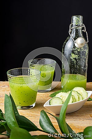 Top view of two glasses with cucumber juice and bottle, bowl with slices, mint and unfocused green branch, on wooden table and bla Stock Photo