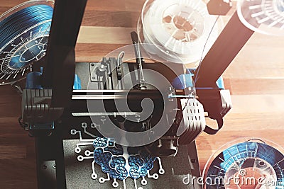 Top view on work space with 3d printer making a symbolic brain sculpture. Wooden desk with filament rolls in warm light. Stock Photo