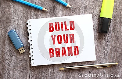 Top view of the workspace. Notepad with BUILD YOUR BRAND text, pen, pencils, flash drive on gray wooden table. Stock Photo