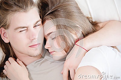 A young man and woman lie in a white bed in a hug and the man slyly looks into the camera while the woman sleeps. The Stock Photo