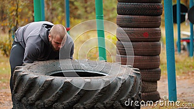 Tough tattooed man bodybuilder in gloves pushes over the truck tire on the ground - training in the autumn forest Stock Photo