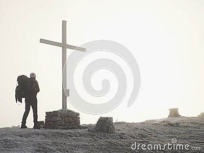 Tourist with big backpack stand at cross memorial on mountain peak. Man is watching into misty Alpine valley bellow. Stock Photo