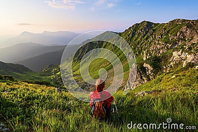 The tourist girl with back sack and tracking sticks sits on the lawn. Relaxation. Mountain landscapes. Wonderful summer day. Stock Photo