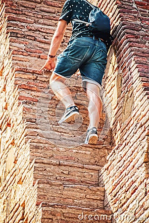 Tourist in jeans shorts and teeshirt with small backpack climbs steep narrow steps in ancient ruin - cropped Stock Photo