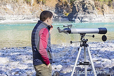 Tourist looks thoughtfully into distance next to telescope at mountain river background in mountains Stock Photo