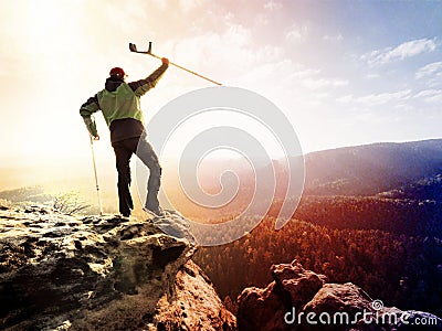 Tourist with medicine crutch above head achieve mountain peak. Hurt backpacker with broken leg in immobilizer stay above valley Stock Photo