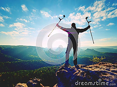 Tourist with medicine crutch above head achieve mountain peak. Hurt backpacker with broken leg Stock Photo
