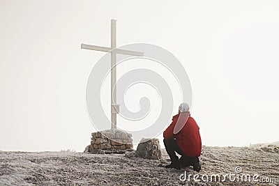 Tourist in red is kneeling at cross memorial on mountain peak. Man is watching into misty Alpine valley bellow. Stock Photo