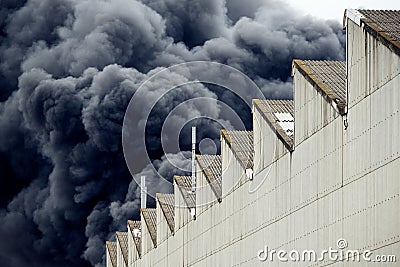 Black plumes of smoke from an accidental toxic industrial fire as seen from a behind a factory building. Stock Photo