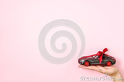 Toy car model tied with a red ribbon and bow on human hand on pink background. Auto dealership and rental, car as gift or present Stock Photo