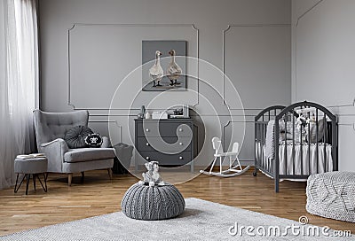 Toy on grey pouf in the middle of trendy baby bedroom with grey wooden cradle, chest of drawers and comfortable armchair, real Stock Photo