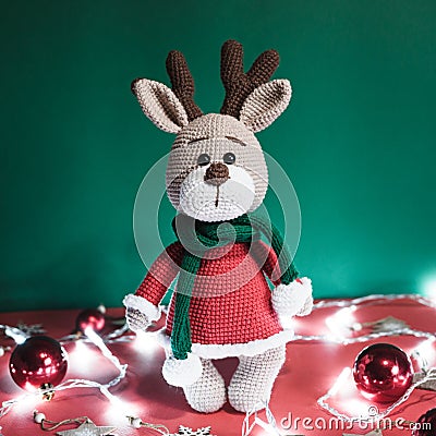 Toy small Christmas deer on a red and green background with decorations and lights from a garland. Cute handmade knitted deer in a Stock Photo