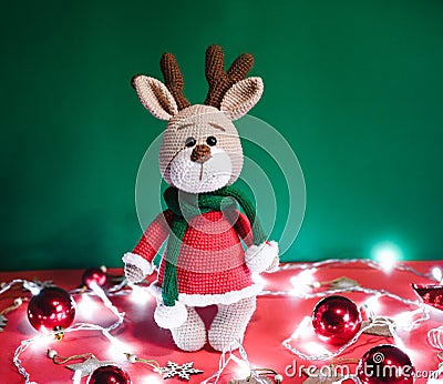 Toy small Christmas deer on a red and green background with decorations and lights from a garland. Cute handmade knitted deer in a Stock Photo