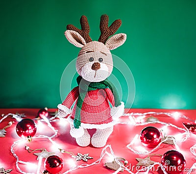 Toy small Christmas deer on a red and green background with decorations and lights from a garland. Cute handmade knitted deer in a Stock Photo