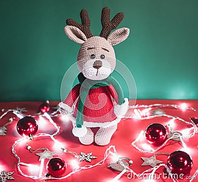 Toy small Christmas deer on a red and green background with decorations and lights from a garland. Cute handmade knitted deer in a Stock Photo