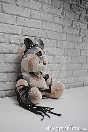 Toy Teddy bear dressed in leather belts and mask accessory for BDSM games on a light background texture of a brick wall Stock Photo