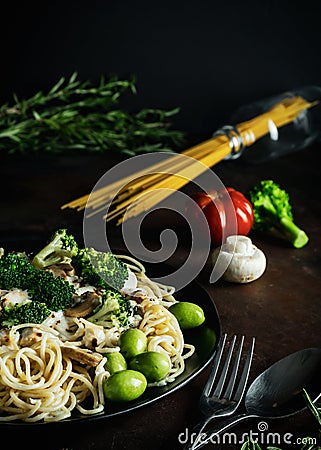 Traditional delicious pasta with chicken, mushrooms, broccoli, cream sauce and green olives on black plate and uncooked spagetti Stock Photo