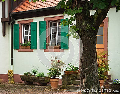 A traditional german house with white walls, green shutters and tile roof, a yard with flower pots and a tree near it Stock Photo
