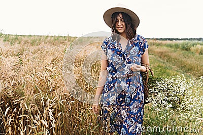 Tranquil summer in countryside. Stylish young woman in blue vintage dress and hat walking with white wildflowers in straw basket Stock Photo