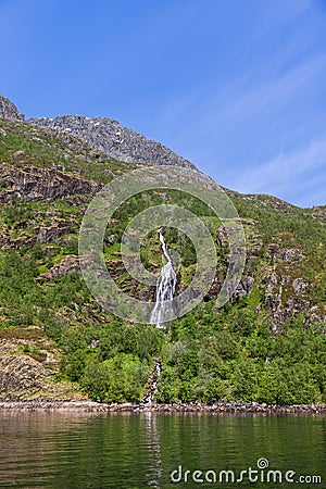 The tranquil waters of Trollfjorden reflect the sunlit, verdant landscape and a thin waterfall descending Stock Photo