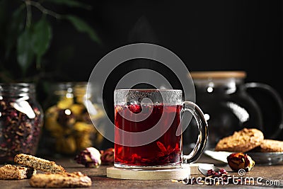 Transparent mug with hot red tea on a dark background, near cookies and tea leaves Stock Photo