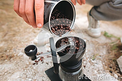 Traveler filling grinder with fresh coffee beans on cliff at lake, preparing for brewing alternative coffee at camping. Making hot Stock Photo