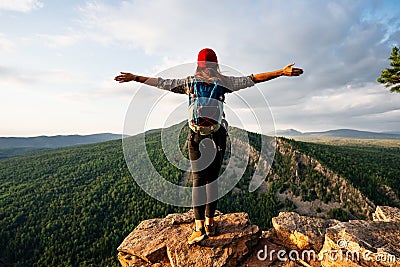 A traveler girl with a backpack is standing on the edge of the mountain, a rear view. Stock Photo