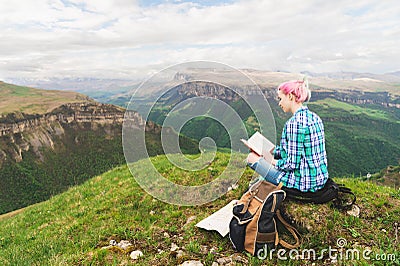 A traveler girl sits in the mountains on the grass and reads a book on the background of epic mountains. The concept of Stock Photo