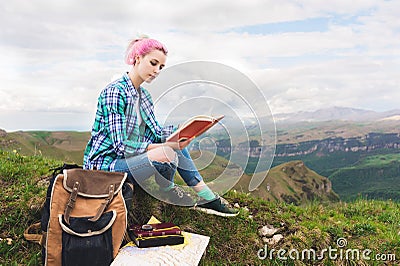 A traveler girl sits in the mountains on the grass and reads a book on the background of epic mountains. The concept of Stock Photo