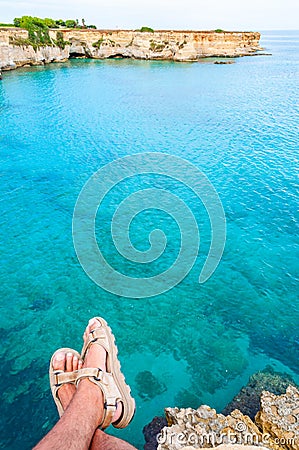 Sitting on the edge of a cliff with dangling legs in travel sandals looking on crystal clear water on Torre Sant Andrea beach with Stock Photo