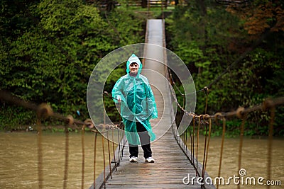 Traveller senior beautiful woman in blue rain jacket cross river by hinged bridge in forest, enjoying silence Stock Photo