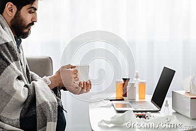 Treating flu and colds at home. Arab guy having fever wrapped in blanket and drinking hot tea, watching film on laptop Stock Photo