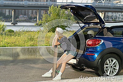 Traveler female relax leaning on longboard at car trunk. Woman resting after skateboarding outdoor Stock Photo