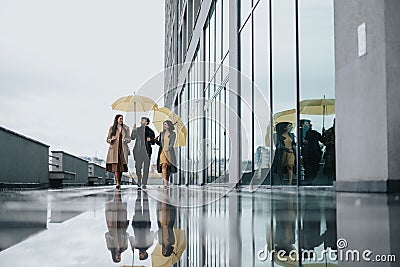 Trio of business partners with yellow umbrellas walking in rainy urban setting. Stock Photo