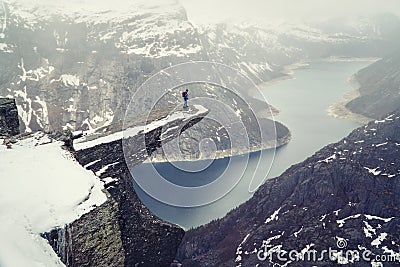 Trolltunga cliff under snow in Norway. Scenic Landscape. Man traveller standing on edge of rock and looking down. Travel Stock Photo