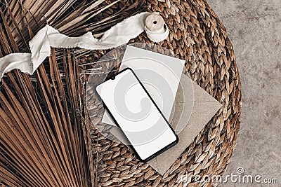 Tropical summer mockups. Black cell phone with empty screen, blank greeting card, envelope and ribbon placed on rattan Stock Photo
