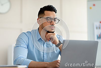 Troubled Arab office worker sitting at table, looking at laptop screen, thinking over business problem at office Stock Photo