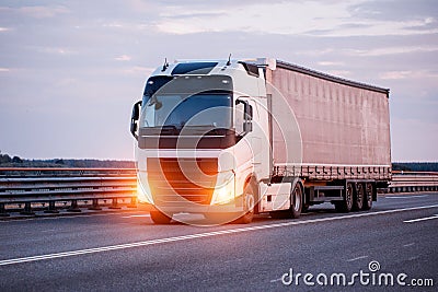 A truck with a semi-trailer curtain transports a load of auto parts in the evening along the highway with headlights on Stock Photo