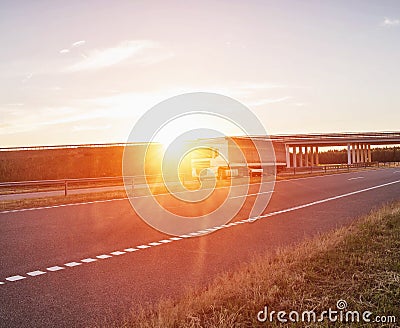 A trucker driver transports cargo on a white wagon against the background of a road bridge and a sunset. Shipping, business and Stock Photo