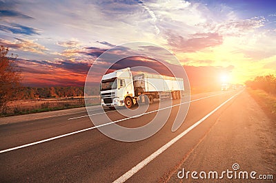 Trucks with a trailers and cars on the countryside road against sky with sunset Stock Photo