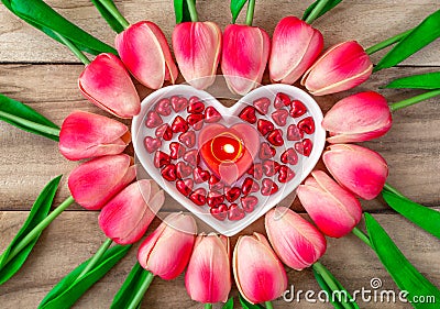 Tulip flowers are laid out in the shape of a heart on a wooden background, among them are a heart-shaped plate and a burning heart Stock Photo