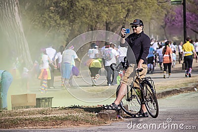 Man on bicycle wearing baseball cap and cargo shorts & sunglasses pauses to take phone pic of participants in Editorial Stock Photo