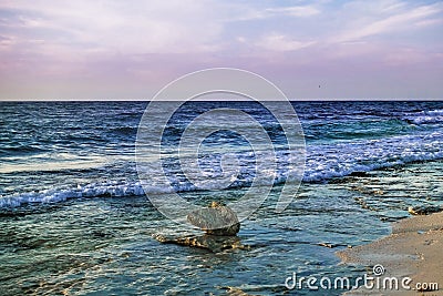 Turquoise waves roll onto the sandy beach. The sun illuminates a bizarre stone on the shore. Stock Photo