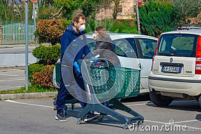 A young coucasian male with white face mask strolling in parking lot with shopping cart. Buying groceries in pandemic in Italy Editorial Stock Photo