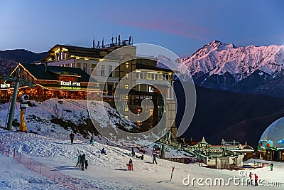 Twilight townscape of Gorky Gorod mountain ski resort on snowy Chugush mountain peak background. People on snowy slope at night sk Editorial Stock Photo